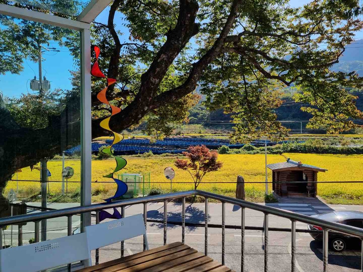 A view from a terrace displays a vibrant landscape, featuring lush greenery and fields of golden rice under a clear blue sky. The scene is complemented by a colorful sculpture hanging from a tree branch, adding a touch of artistic flair.