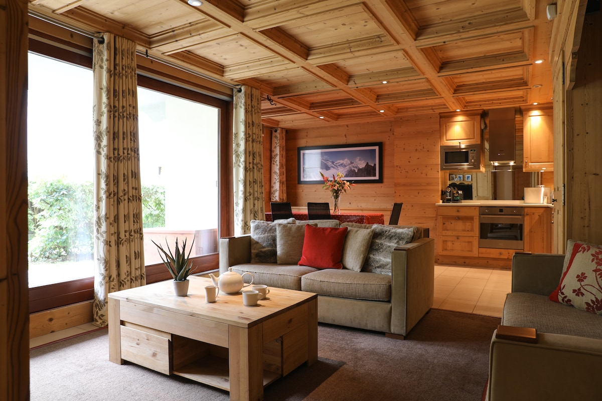 The lounge area features wooden paneling and a coffered ceiling, complemented by natural light from large windows. A spacious sofa with colorful cushions surrounds a wooden coffee table, while a dining table is visible in the background. Modern kitchen appliances are integrated along one wall.