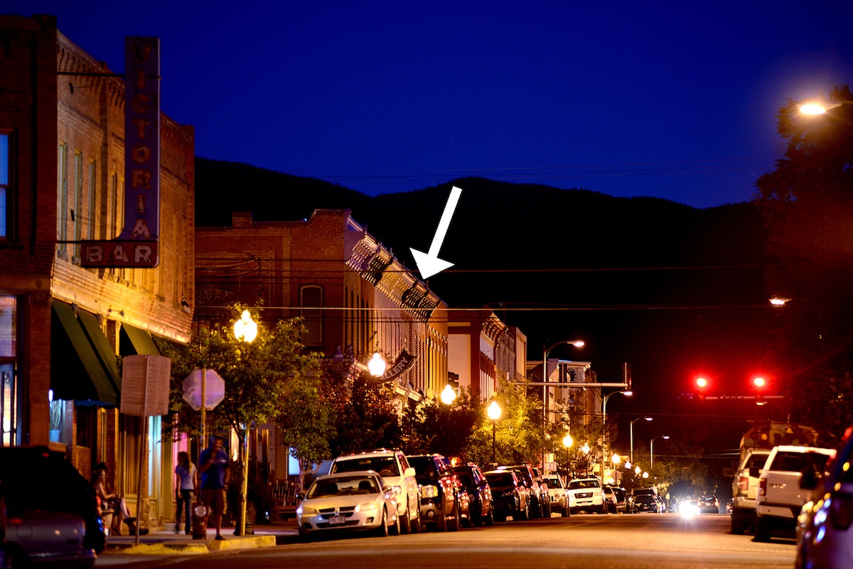 An evening view of a historic downtown street features buildings lined with warm lighting and parked cars. The serene atmosphere is enhanced by silhouette mountains in the background, while street lamps illuminate the sidewalks, guiding the way along the bustling main street.