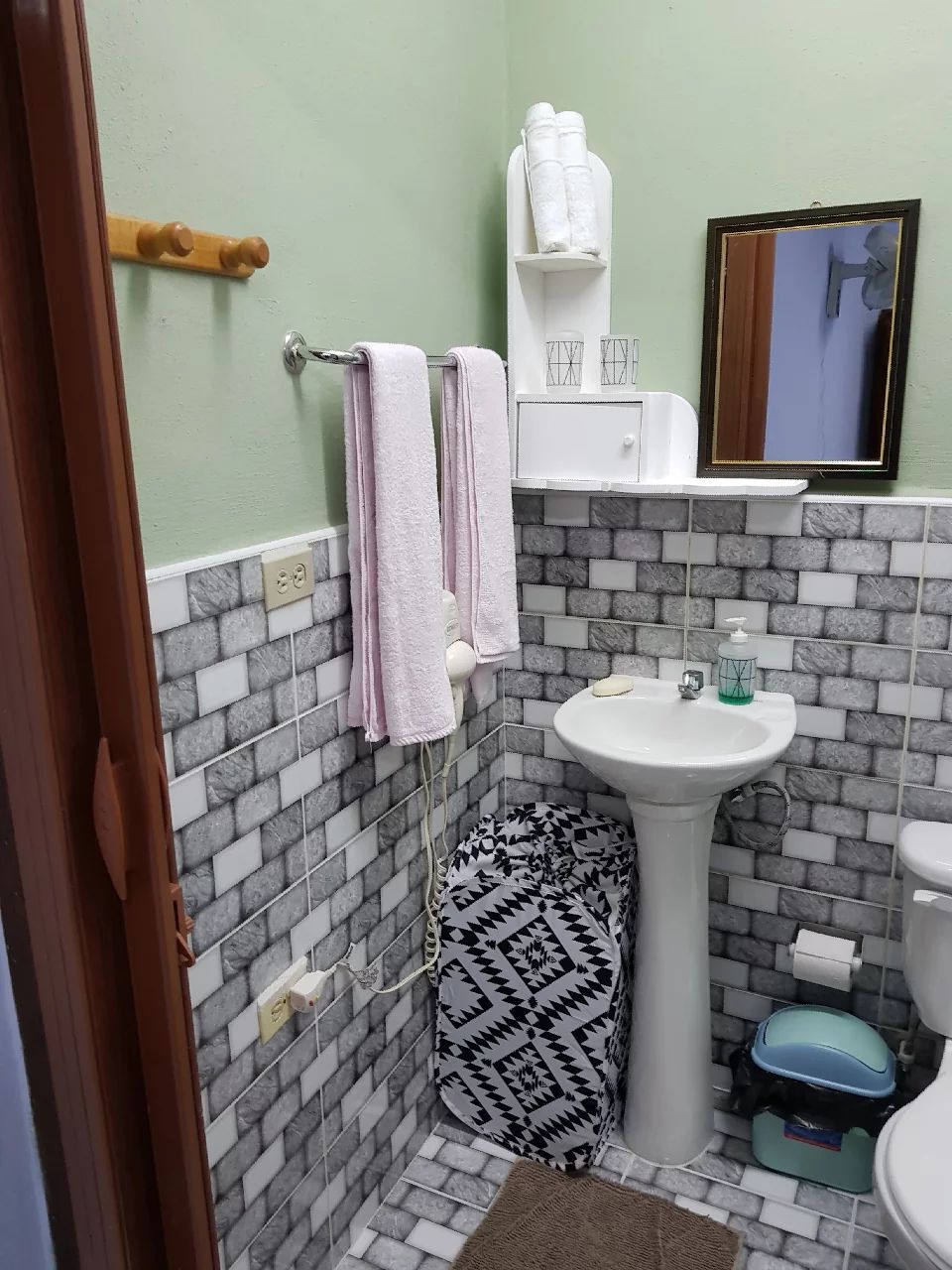 A compact bathroom features a pedestal sink with a mirror above it. Soft towels hang on a rack beside the sink, complemented by a patterned mat on the floor. The wall displays stylish mosaic tiles in light and dark tones, creating a modern look.