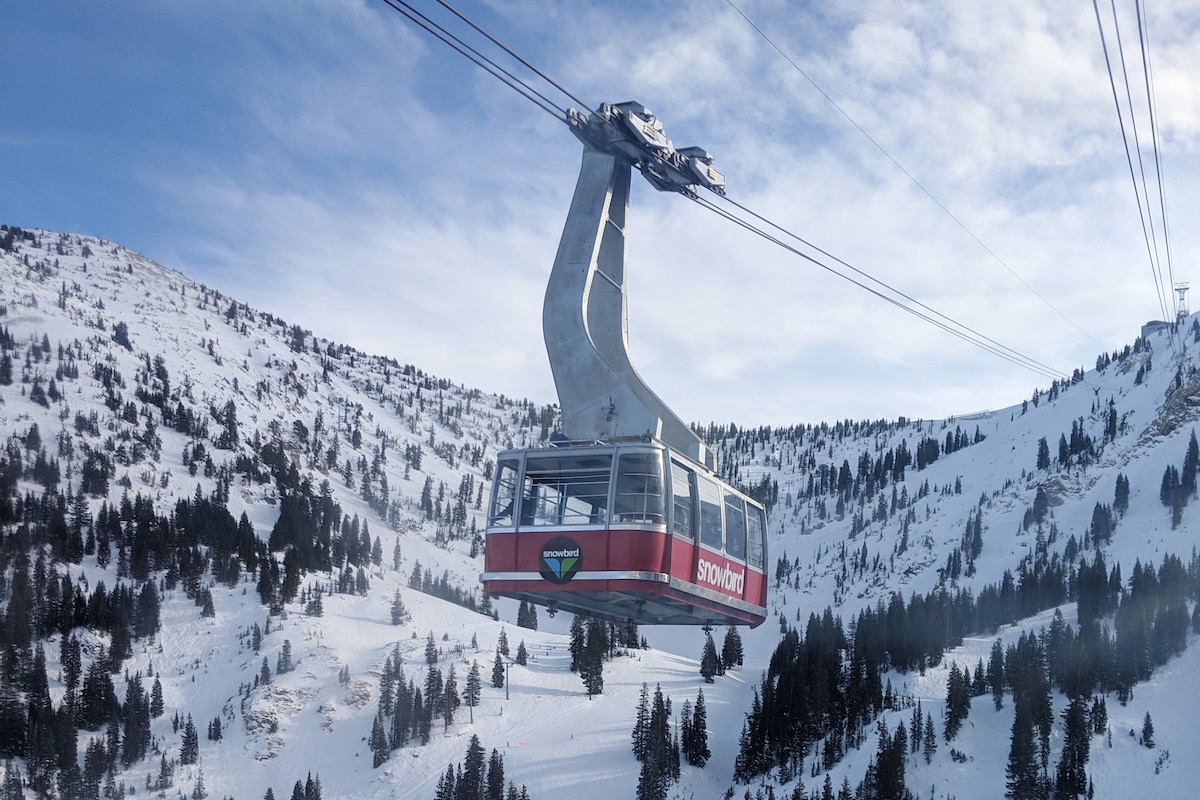 A gondola cabin is suspended above a snow-covered landscape, with steep mountain slopes and evergreen trees visible below. The clear blue sky enhances the scene, showcasing the beauty of the winter environment.