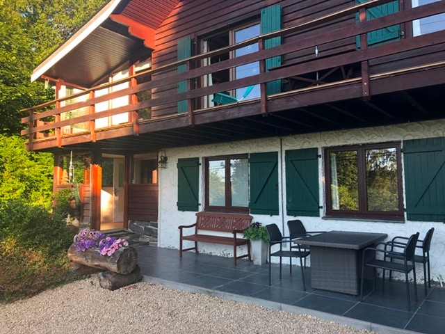 The exterior of a wooden chalet is presented, showcasing a covered terrace with a dining table and chairs. Green shutters frame the windows, and colorful flowers decorate the entrance. A gravel area surrounds the property, enhancing the natural setting.