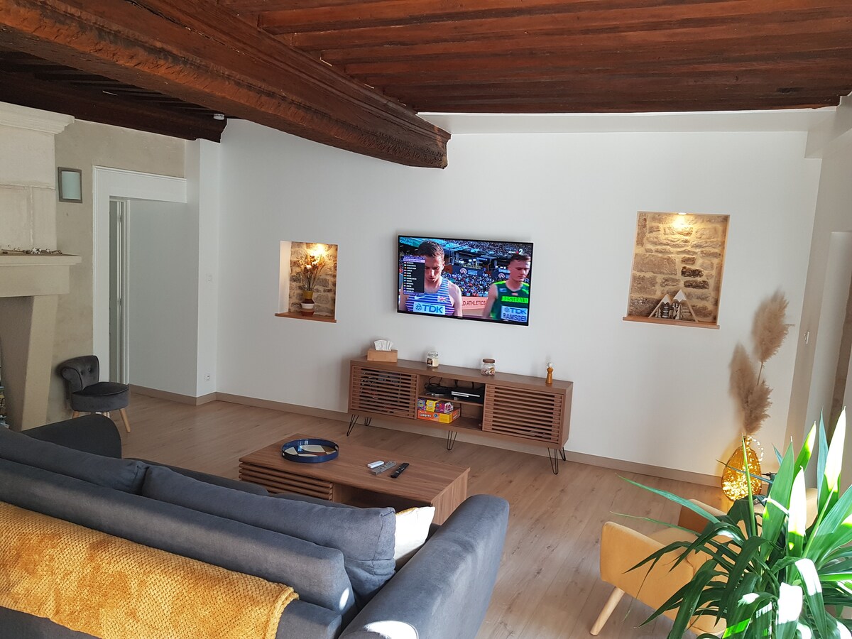 A welcoming living area is highlighted by a spacious sofa facing a large TV mounted on a white wall. Natural light filters through windows, illuminating decorative wall niches. The wooden ceiling beams add character, while a green plant brings a touch of nature to the setting.