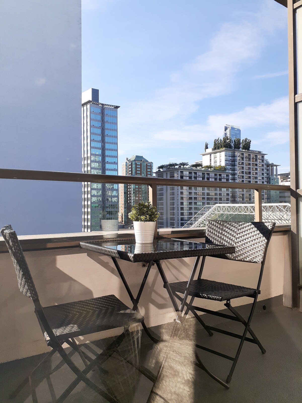 A small balcony offers a bistro table and two folding chairs, with a potted plant centerpiecing the setup. The urban skyline is visible in the background, showcasing a mix of modern high-rise buildings against a clear blue sky.