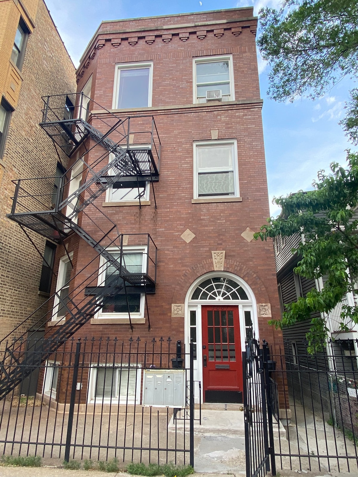 A red brick four-flat building is captured from street level, showcasing an ornate entrance framed by a decorative arch. Metal fire escapes are visible on either side, complementing the exterior while large windows allow light to enter. A black wrought iron fence lines the front property.