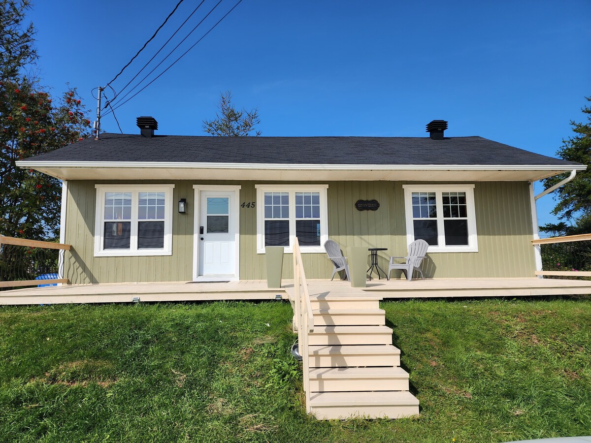 The exterior of the house features a light green facade with white trim. A set of stairs leads up to the front door, flanked by two chairs on a spacious porch. Large windows allow for ample natural light, complemented by a clear blue sky.