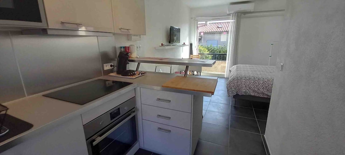 A modern kitchen is seen featuring sleek white cabinetry and a wood countertop. The room opens up to a bright living area with a view of a small outdoor space, while a comfortable sleeping area is indicated by a bed with simple bedding in the background.