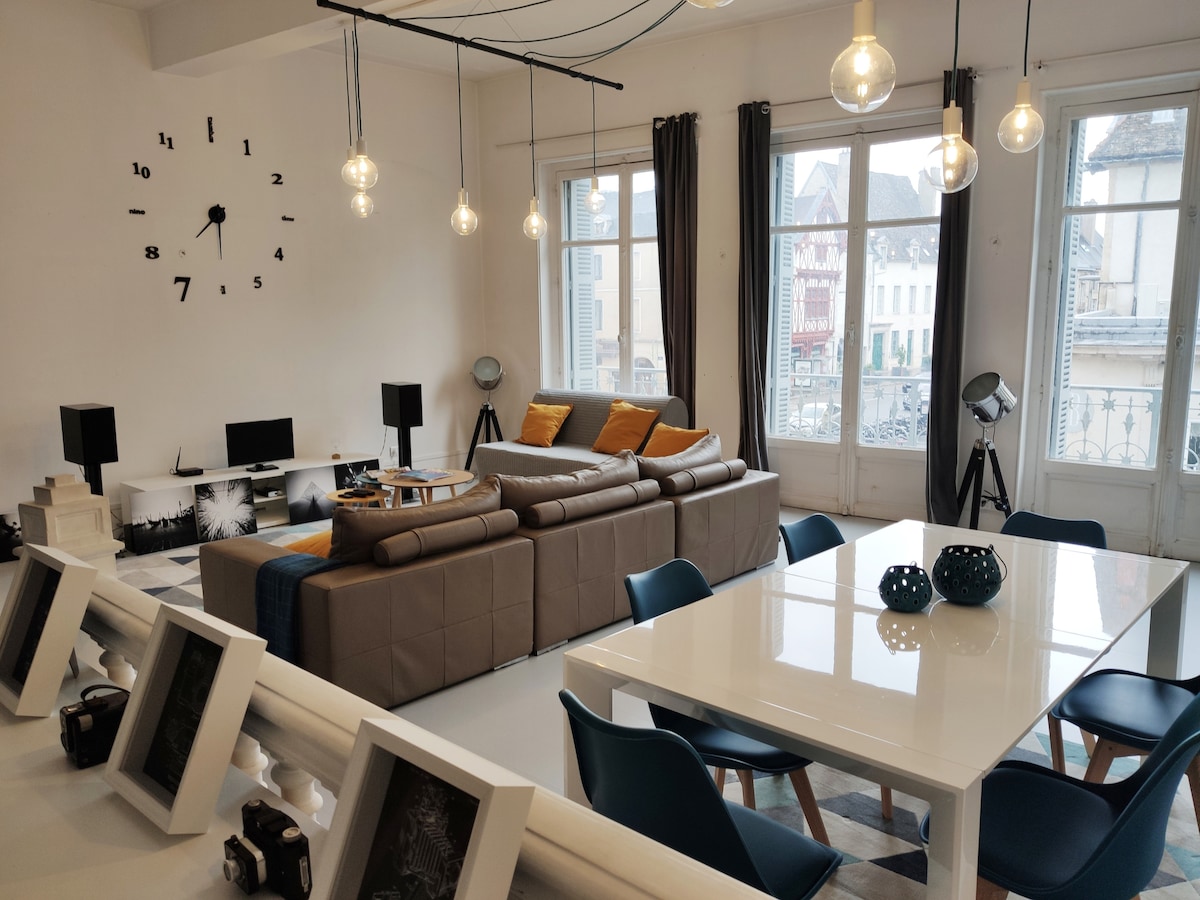 A bright living area is presented, featuring a comfortable sectional sofa with decorative cushions. A large dining table is accompanied by blue chairs, while a clock adorns the wall. Large windows allow natural light to fill the space, complemented by stylish pendant lights.
