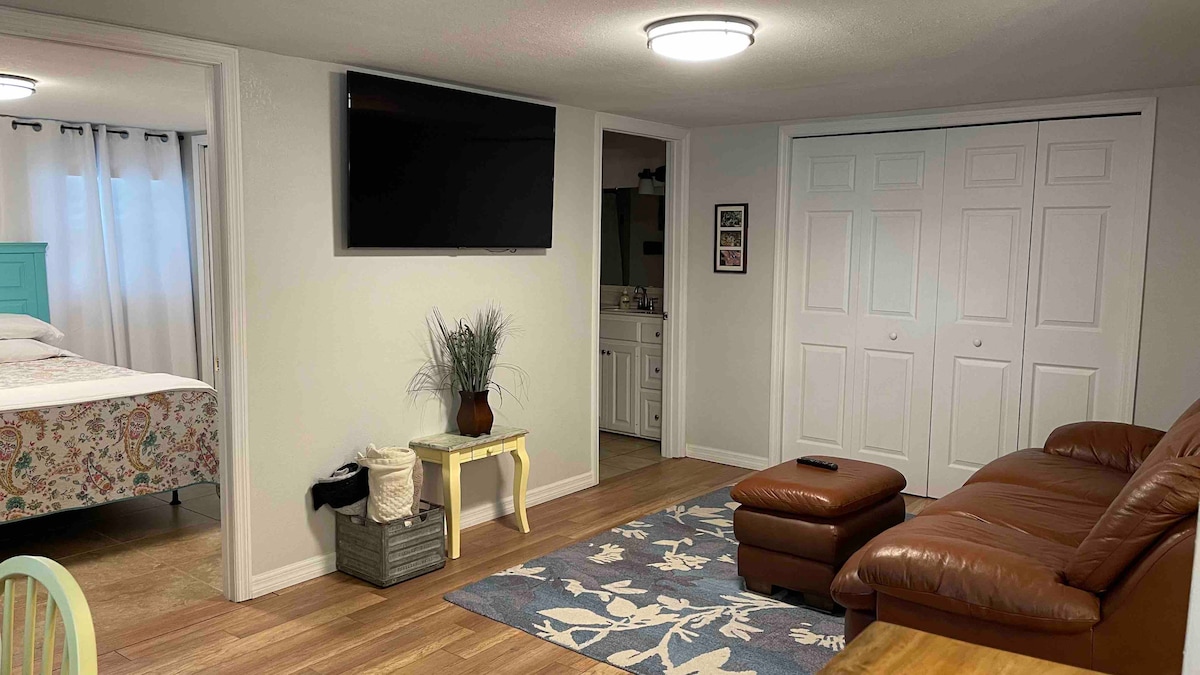 A cozy living area features a brown leather sofa, a patterned area rug, and a small yellow accent table. A flat-screen television is mounted on a light wall. Dual closets are visible in the background, along with a glimpse of the adjoining sleeping space.
