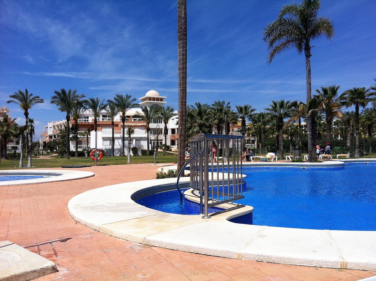 A spacious outdoor area features inviting swimming pools surrounded by palm trees and lush gardens. Sun loungers are placed around the pool area, while a gentle incline leads to a blue water feature. Colorful buildings can be seen in the background under a clear sky.