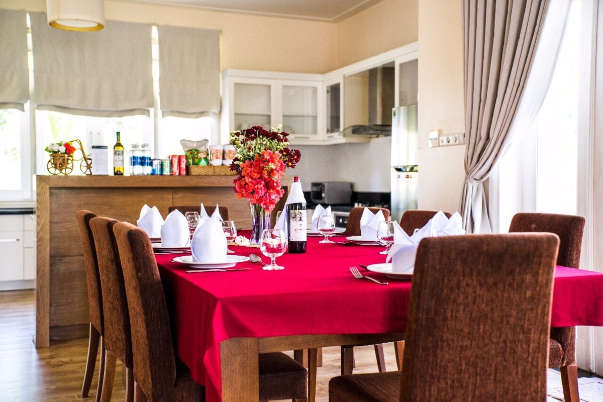 A spacious dining area is presented with a red tablecloth covering a large wooden table set for eight. Neatly arranged white napkins and glassware complement the setting. In the background, an equipped kitchen is visible, featuring glass-front cabinets and various pantry items.
