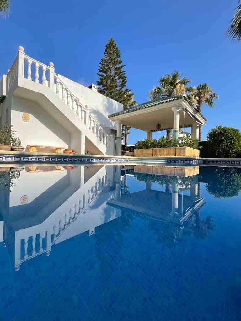 A clear blue swimming pool reflects the surrounding villa and covered pergola. The structure features white walls and decorative elements, accompanied by lush greenery. A staircase leads to an upper level, enhancing the tranquil outdoor setting under a bright blue sky.