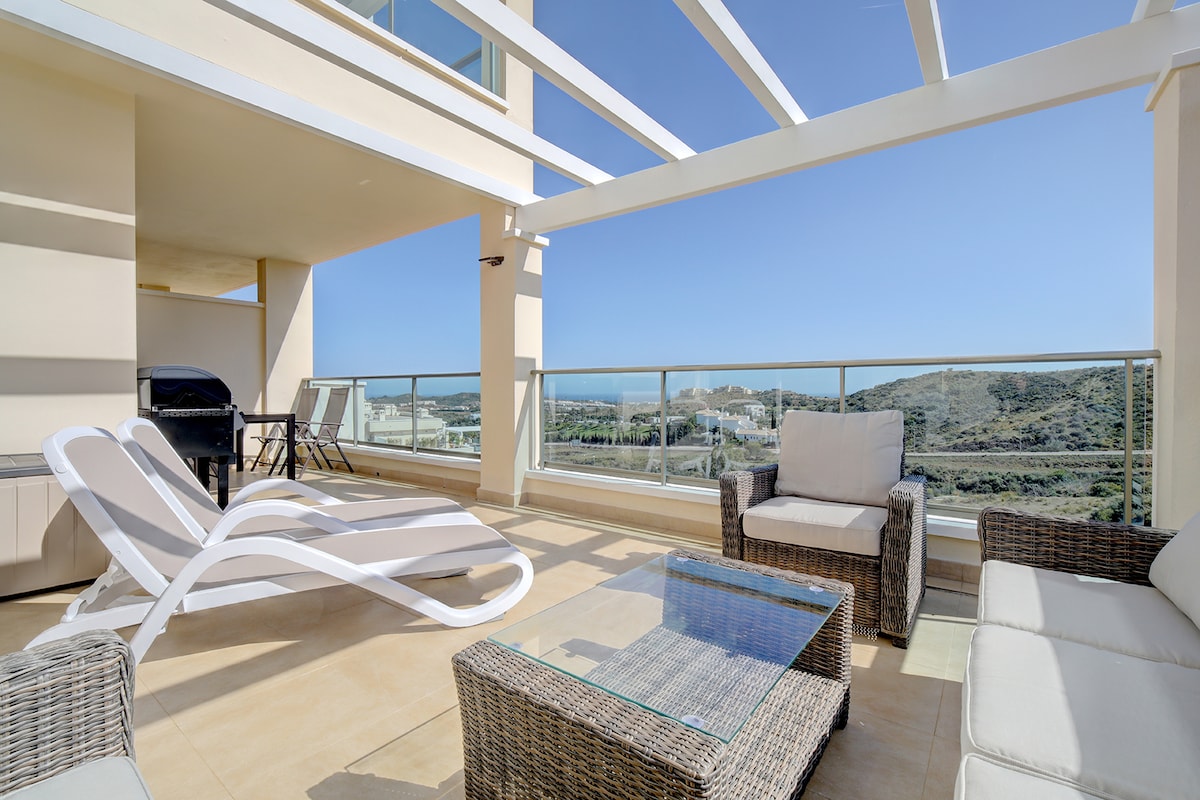 An airy terrace features modern outdoor furniture, including a lounge chair and seating area with cushions. A glass-top table is positioned at the center, and a barbecue grill is located in the background, with expansive views of the sea and mountains visible beyond the glass railing.