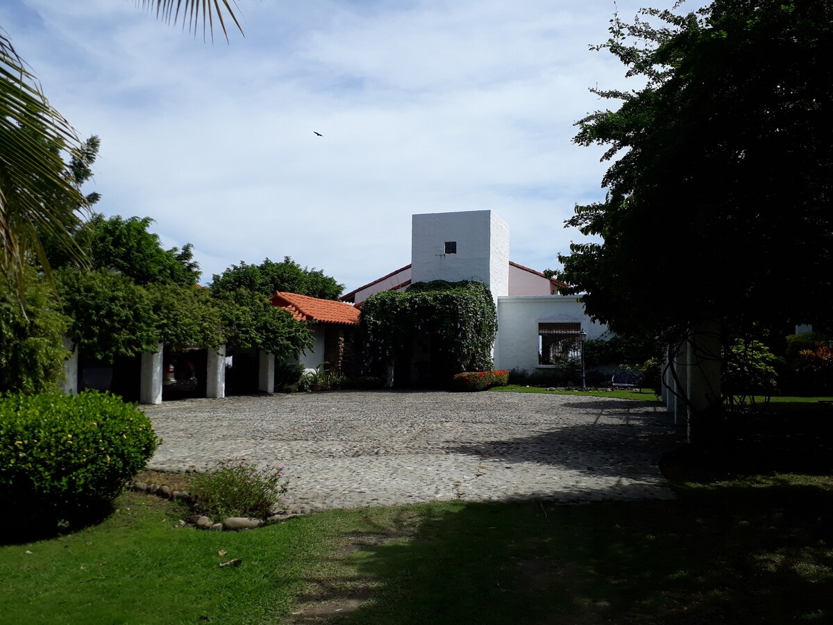 The exterior of the property is set within a serene landscape, featuring a paved courtyard surrounded by lush greenery. The building's architecture combines white walls and terracotta roofs, with ample trees offering natural shade throughout the area.