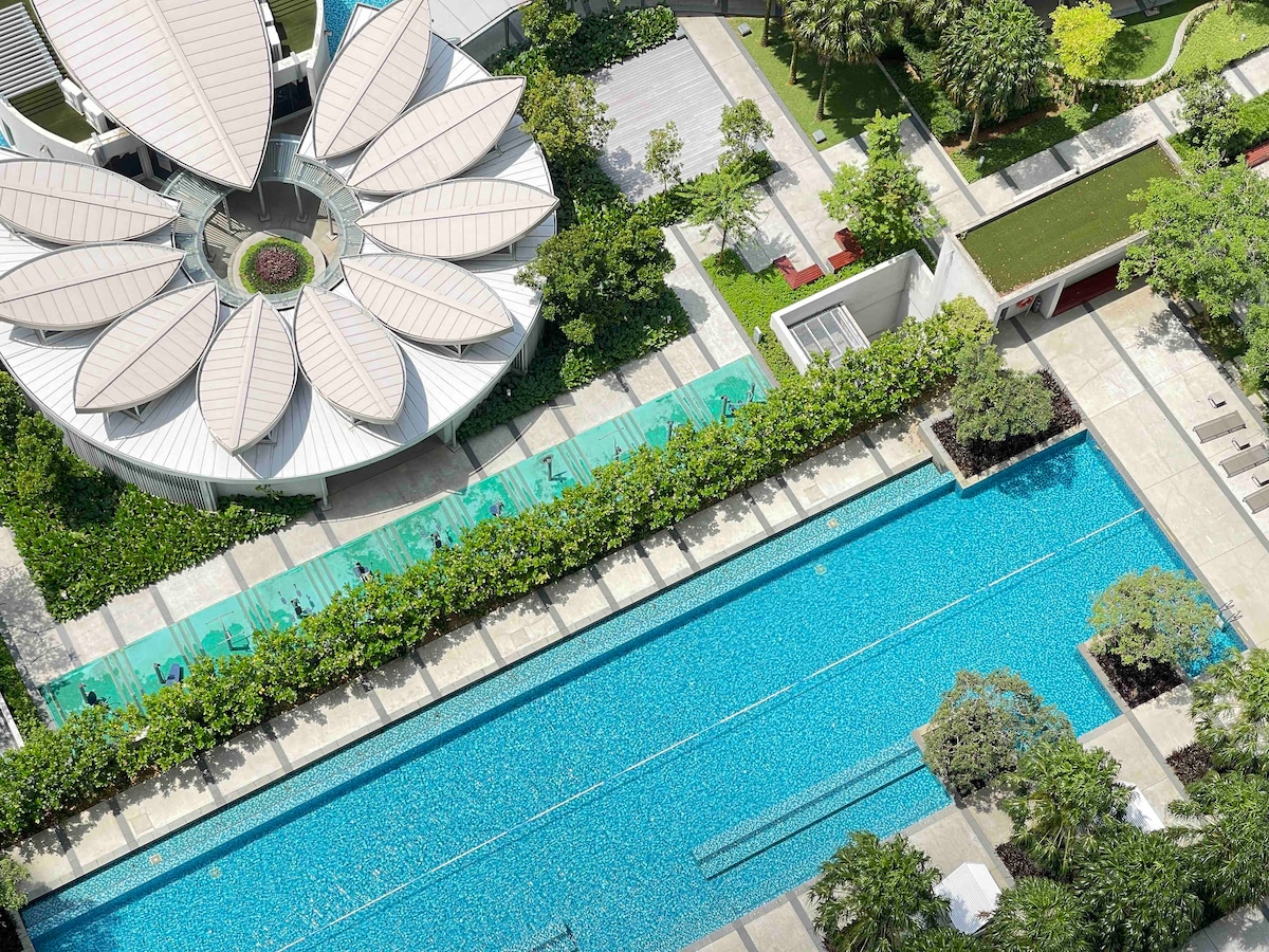 Aerial view of a spacious outdoor area featuring a large swimming pool with clear blue water. Surrounding greenery adds to the serene atmosphere, while modern architectural elements are visible, including a flower-shaped pavilion. Sun loungers line the pool area, inviting relaxation.