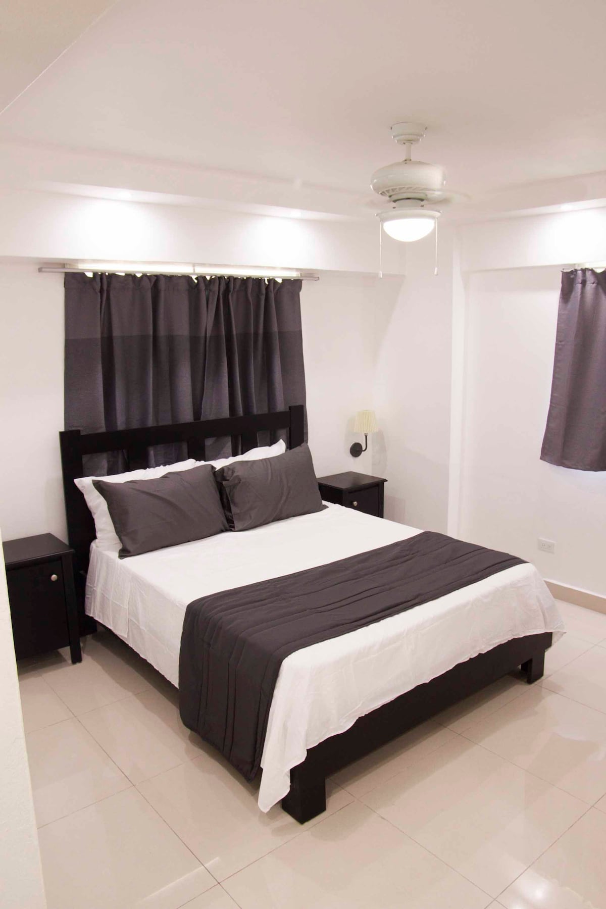 A well-lit bedroom features a spacious bed with gray and white bedding, complemented by matching curtains. Nightstands are positioned on either side of the bed, and a ceiling fan is visible above, providing a comfortable ambiance.
