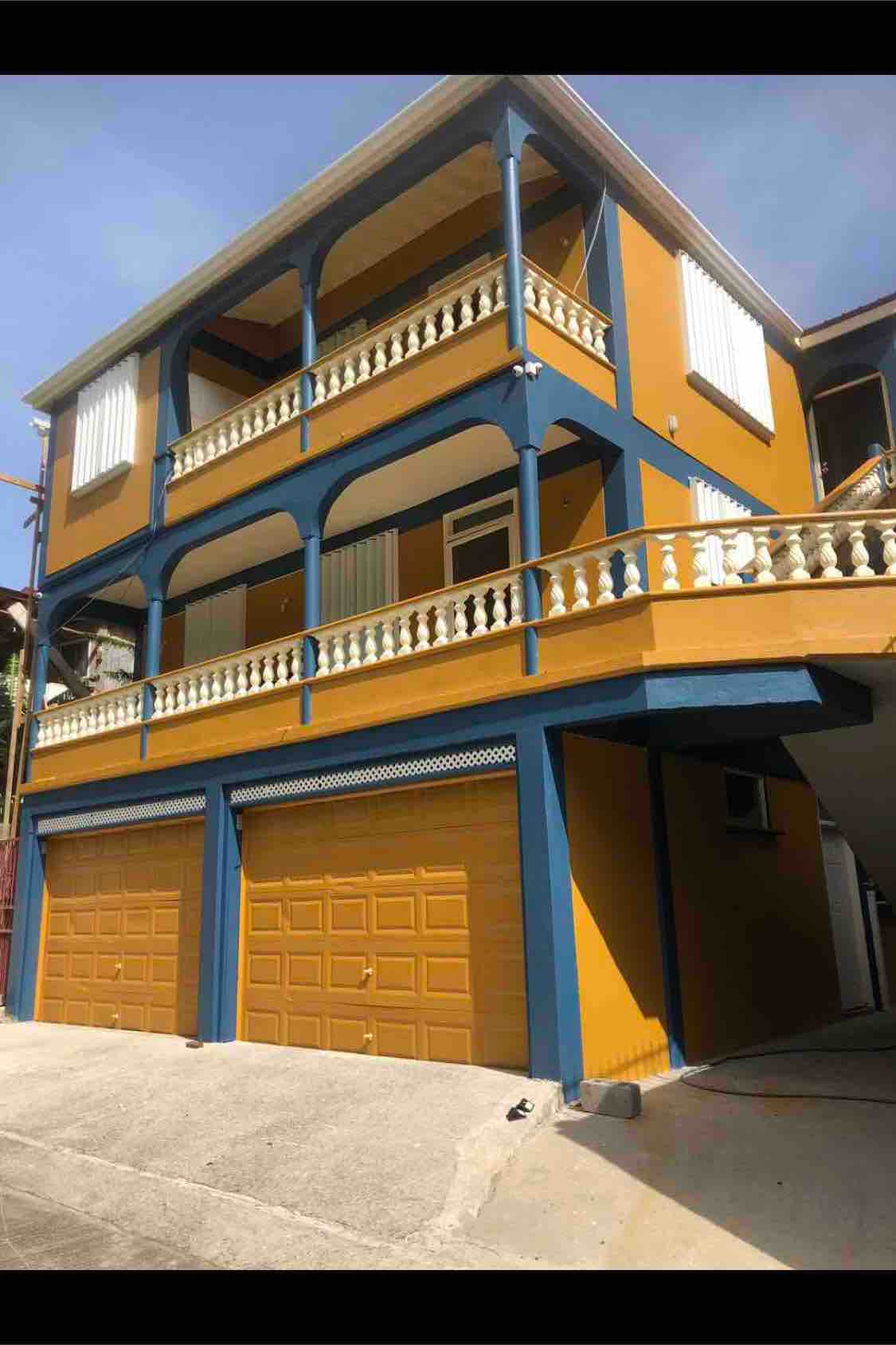 The exterior view features a vibrant building with blue and yellow accents. A spacious balcony with arched supports surrounds the upper floor, while two garage doors are visible at the ground level. The sloped entrance path adds to the structure's unique architectural style.