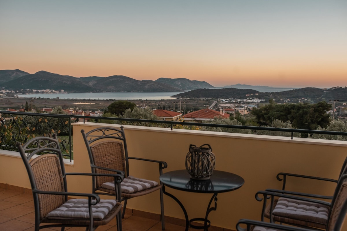 A terrace overlooking a panoramic view features a small round table surrounded by four wicker chairs. The background showcases rolling hills and a tranquil sea under a soft sunset sky, creating a serene outdoor space.