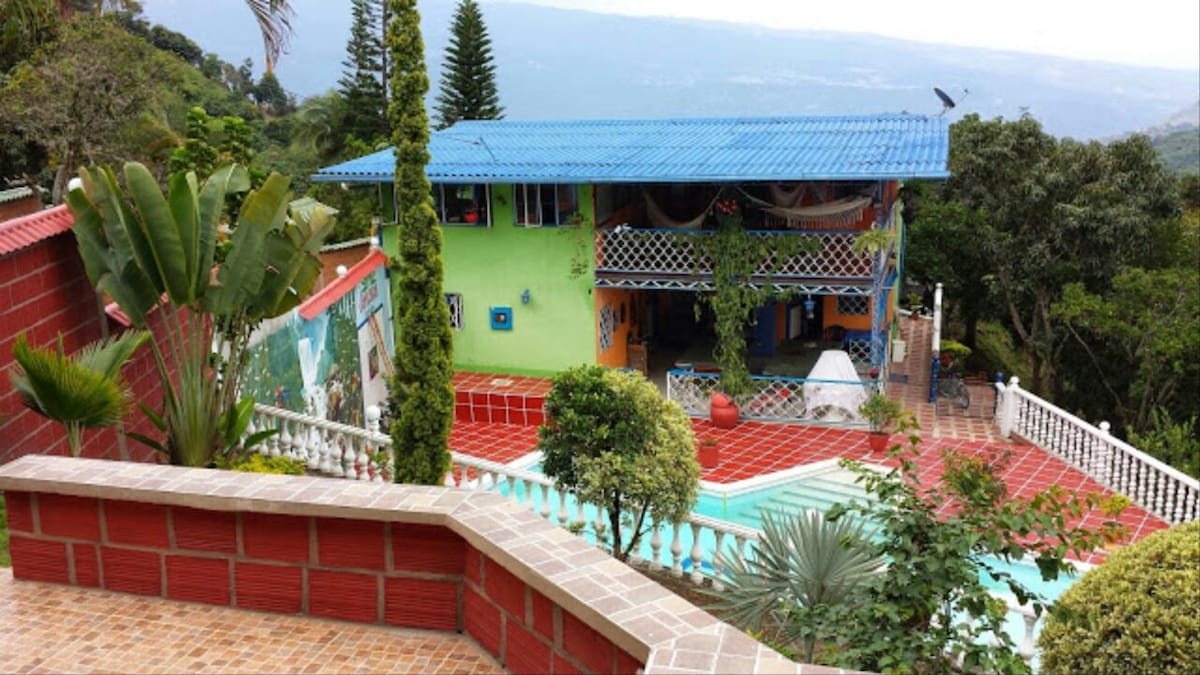 A multi-level property is situated within a lush landscape, featuring vibrant green and orange exteriors. A pool is visible in the foreground, surrounded by a tiled terrace. Tropical plants accentuate the garden, enhancing the overall natural setting.