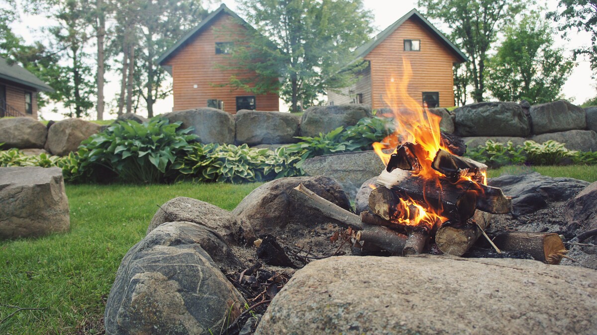 A fire pit surrounded by large rocks is seen with a crackling fire in the center. Green grass and vibrant plants frame the area, while two rustic cabins are visible in the background, partially hidden by trees.