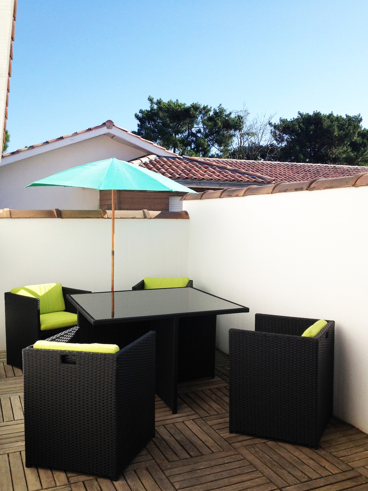 A private terrace is featured, furnished with a square table surrounded by four chairs. An umbrella offers shade, while a wooden deck adds warmth to the space. Blue skies and lush trees are visible in the background, enhancing the tranquil outdoor setting.