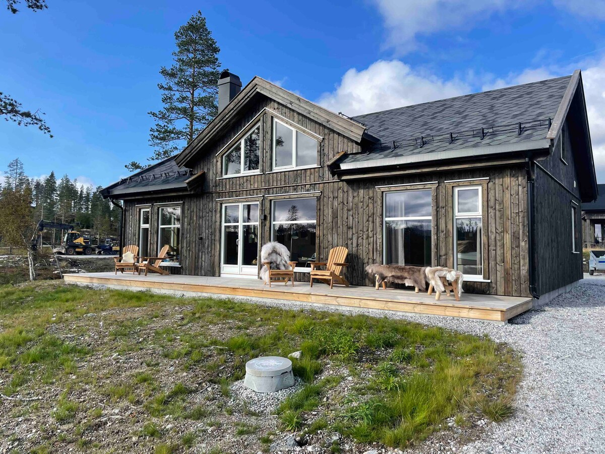 The exterior of a modern cabin is shown, featuring a wooden facade with large windows. Two wooden chairs are positioned on a deck, and a small dog is seen nearby. The setting includes grassy areas and trees, with a partly cloudy sky in the background.