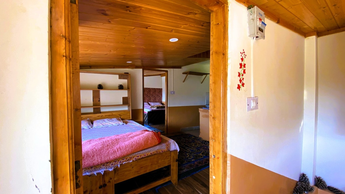 A welcoming interior showcases a wooden frame bed covered with a soft pink blanket. Shelves with decorative items line the wall. A doorway leads to a second room, indicating a cozy, connected living space. Natural light filters in from windows, creating a warm atmosphere.