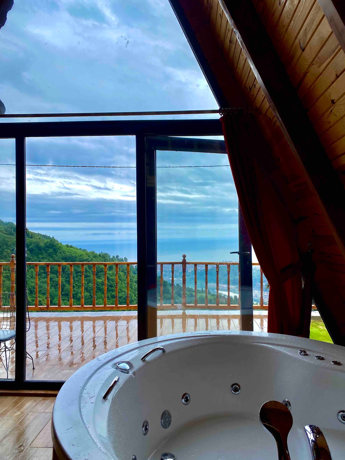 A jetted tub is positioned in the foreground, overlooking a spacious deck. Large glass doors provide an unobstructed view of the sea and surrounding hillside, allowing natural light to fill the area. The combination of wooden beams and modern design creates a harmonious ambiance.