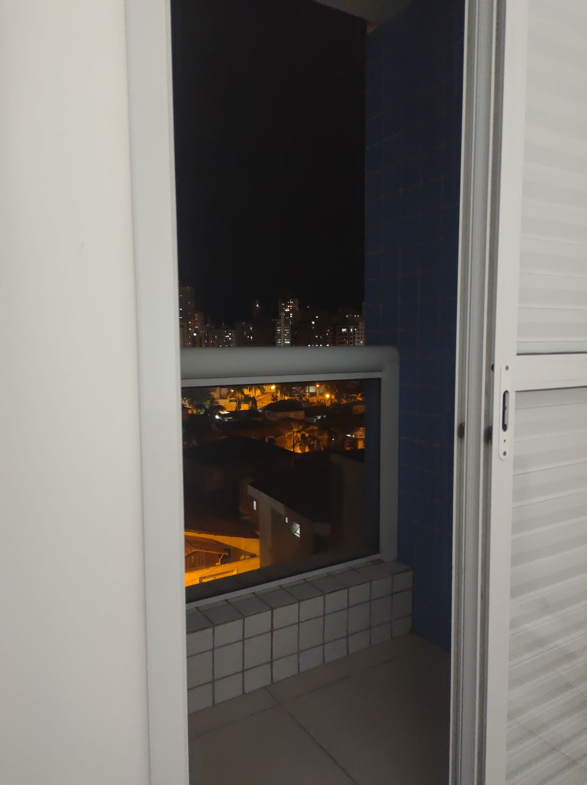 A view from the balcony offers a glimpse of the city at night, with illuminated streets and buildings visible in the distance. The sliding glass door frames the urban landscape, highlighting the contrast between the interior and the vibrant lights outside.