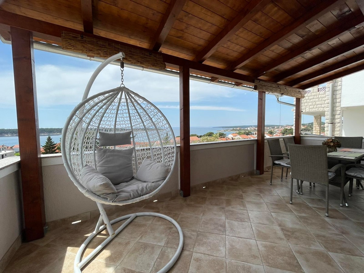 A spacious terrace is seen featuring a hanging chair and a dining area. The overhead wooden beams provide architectural interest. Scenic views of the water and surrounding area are visible in the background, with the soft tones of tiled flooring enhancing the outdoor space.