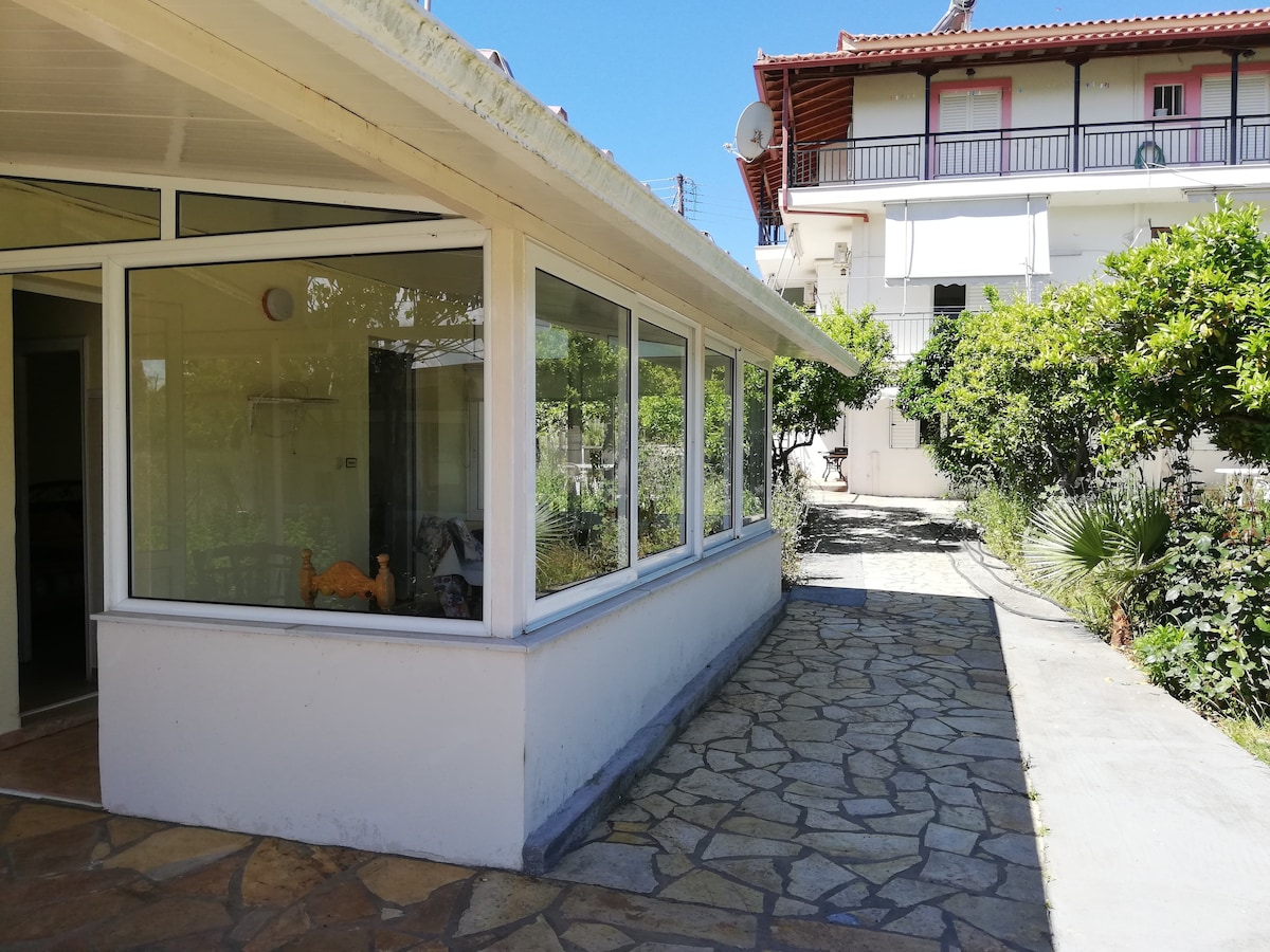 A pathway lined with stone leads to a sunlit area with large glass windows, providing views of the surrounding greenery. A well-kept garden is visible in the background, framed by trees and plants, creating a tranquil outdoor setting.