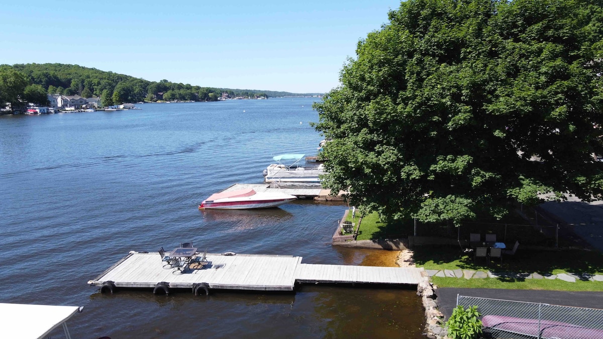 A private dock extends into the calm waters of Lake Hopatcong, with a boat moored alongside. A shaded area under a large tree is visible, accompanied by a small grassy space and a few outdoor chairs. The clear blue sky enhances the tranquil scene.