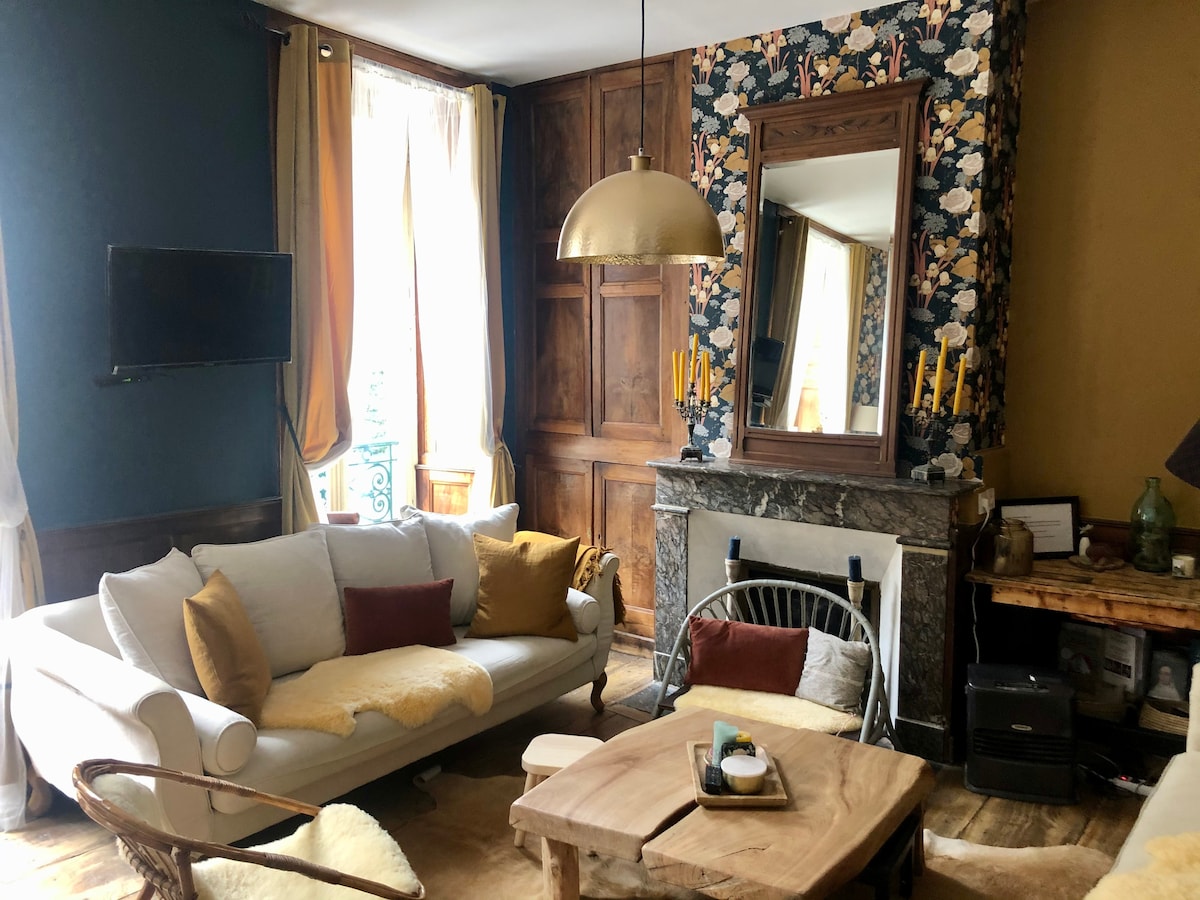 A cozy living room is displayed, featuring a light sofa adorned with colorful cushions. A rustic coffee table is centered in the space, while a decorative mirror reflects soft light. Windows are framed with curtains that add warmth to the environment.