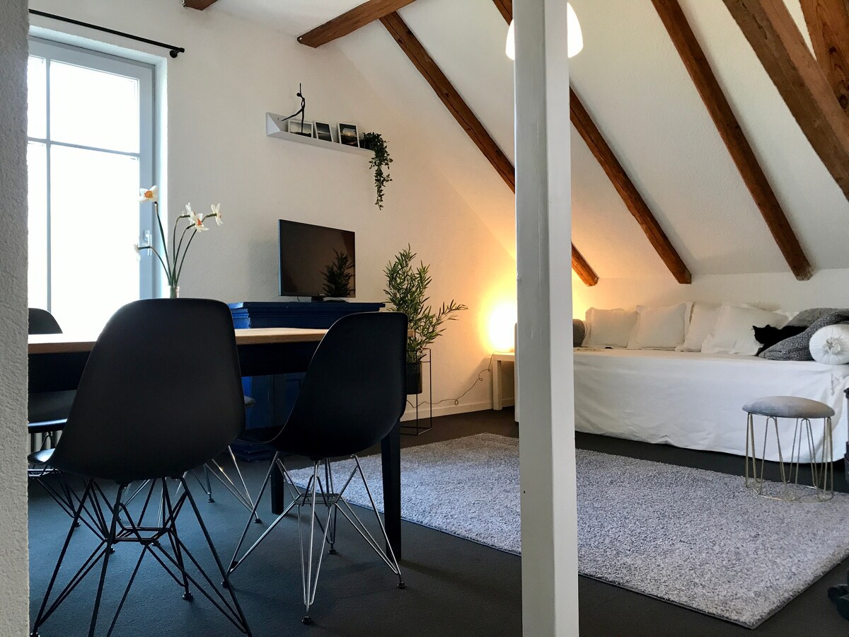 A bright living space features exposed wooden beams and a large window allowing natural light. A dining table with black chairs is positioned near a cozy seating area, which includes a sofa and a small side table. A soft rug adds comfort to the floor.