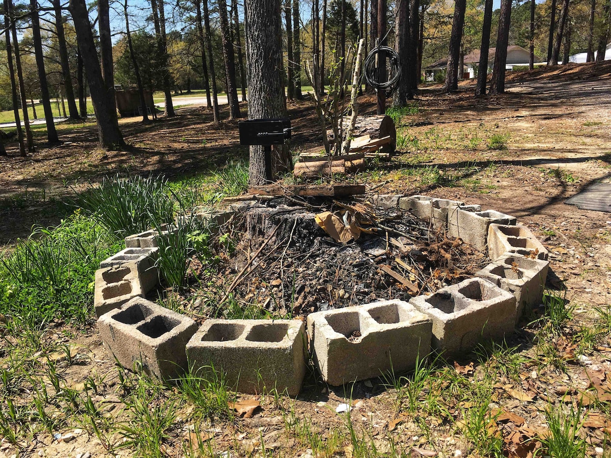 A circular fire pit, surrounded by cinder blocks, is located within a natural setting of grass and scattered leaves. Nearby, trees provide shade and a sense of privacy, creating a suitable space for outdoor gatherings. The area appears well-maintained and inviting for evening fires.