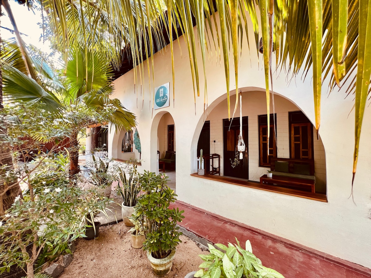The facade of a boutique hostel features arched windows and a sandy pathway bordered by lush greenery. Potted plants are visible, adding a natural element to the inviting entrance. A sign is positioned above the door, indicating the hostel's name.