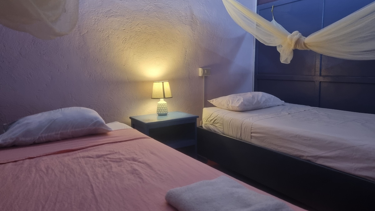 A room featuring two single beds with soft linens, positioned beside a nightstand with a lamp. The walls are painted in a light tone, contributing to a calm ambiance. A gentle glow emanates from the lamp, enhancing the inviting atmosphere.