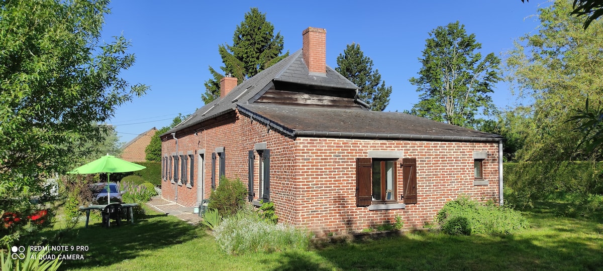 A charming red brick house is situated in a peaceful garden, surrounded by trees and greenery. The exterior features a sloped roof and several small windows. A patio set can be seen in the yard, accompanied by a green umbrella for shade.