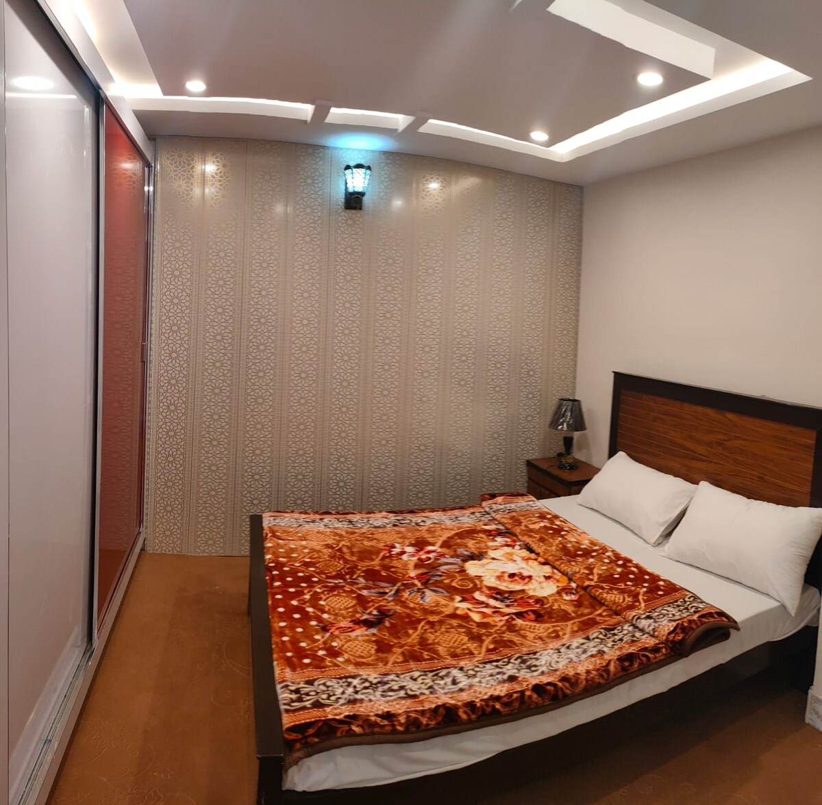 A spacious bedroom features a double bed covered with a decorative blanket. Soft lighting is provided by recessed ceiling lights and a bedside lamp. A large wardrobe is visible along one wall, with a contemporary patterned accent wall enhancing the room's design.