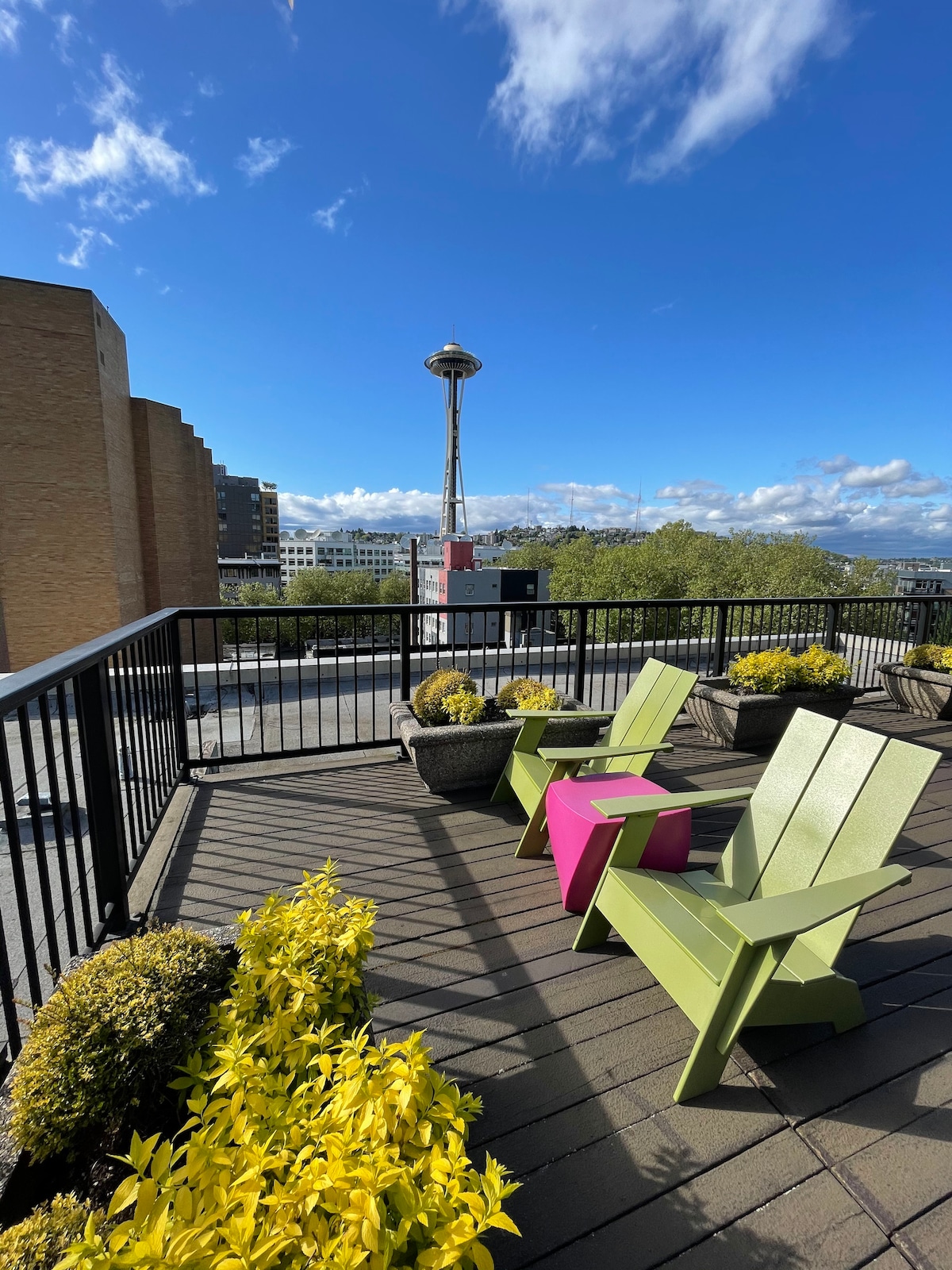 A private balcony features two brightly colored chairs, one in green and the other in pink, surrounded by blooming yellow plants. The Space Needle is prominently visible in the background against a clear blue sky, providing a striking urban view.