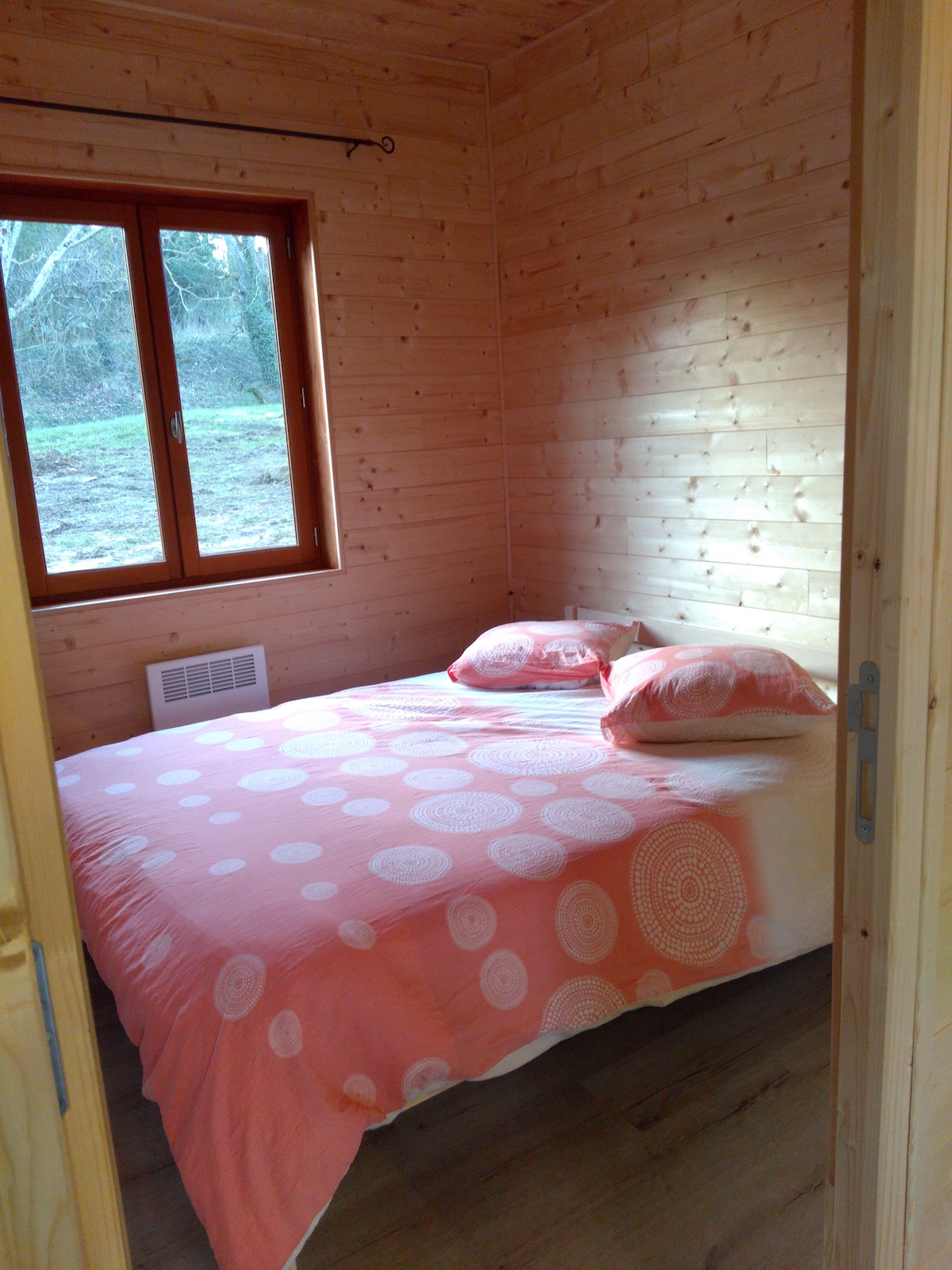 A compact sleeping area is enclosed by wooden walls, featuring a double bed with a coral-colored bedspread adorned with circular patterns. Two matching pillows rest at the head of the bed. A window provides natural light and offers a view of the surrounding greenery.