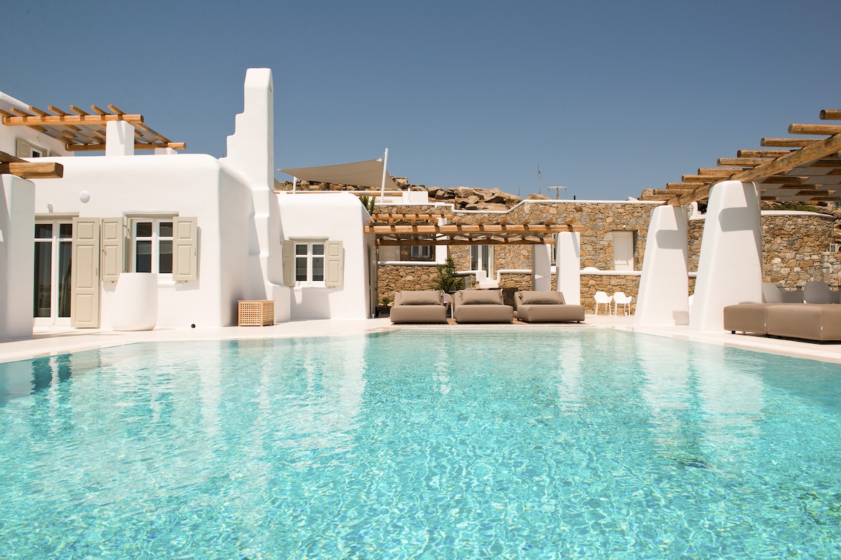 An expansive infinity pool sparkles under the bright sun, surrounded by stylish loungers and shaded by wooden pergolas. The villa's whitewashed architecture features large windows with traditional shutters, reflecting the serene ambiance of Mykonos.