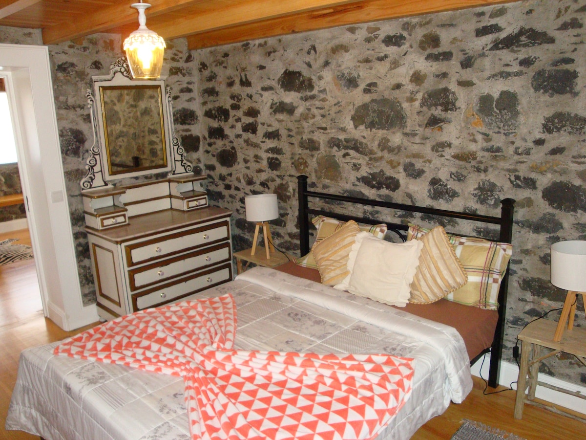 A cozy bedroom features a stone accent wall, highlighting a spacious bed adorned with a patterned blanket and decorative pillows. A stylish mirror is positioned above a dresser, complemented by two bedside lamps on wooden stands, creating a warm and inviting space.