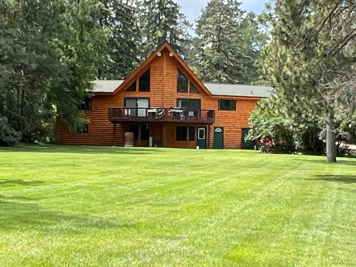 A large, rustic wooden home features a sloped roof and expansive deck, surrounded by lush green grass. Tall trees frame the property, creating a natural backdrop. The home showcases multiple windows, enhancing its inviting character while providing access to outdoor areas.