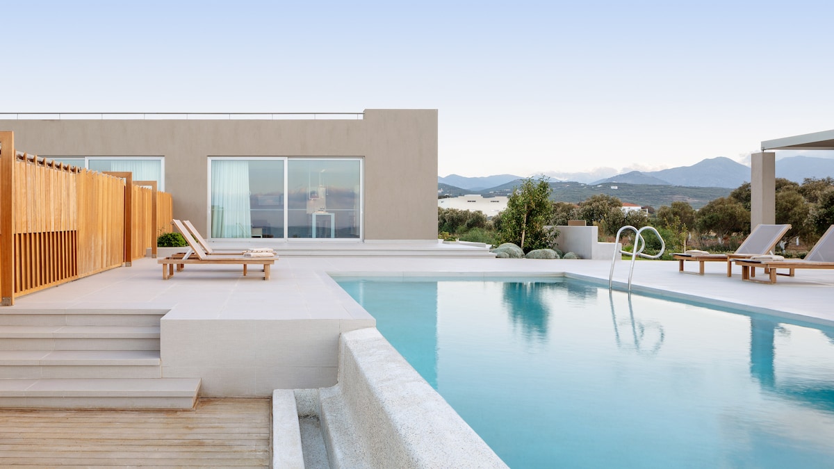An infinity pool reflects the tranquil landscape, surrounded by modern wooden decking and lounge chairs. A sleek villa features large glass doors that open to the outdoor space, seamlessly integrating the indoor and outdoor environments.