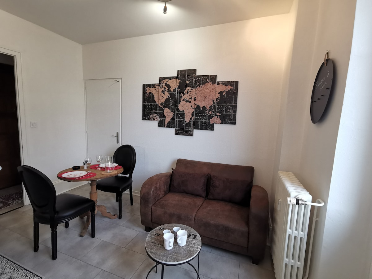 A living area features a brown sofa facing a wall adorned with a world map. A small round table with two black chairs occupies the space, paired with a coffee table. Natural light is allowed in through a nearby doorway.