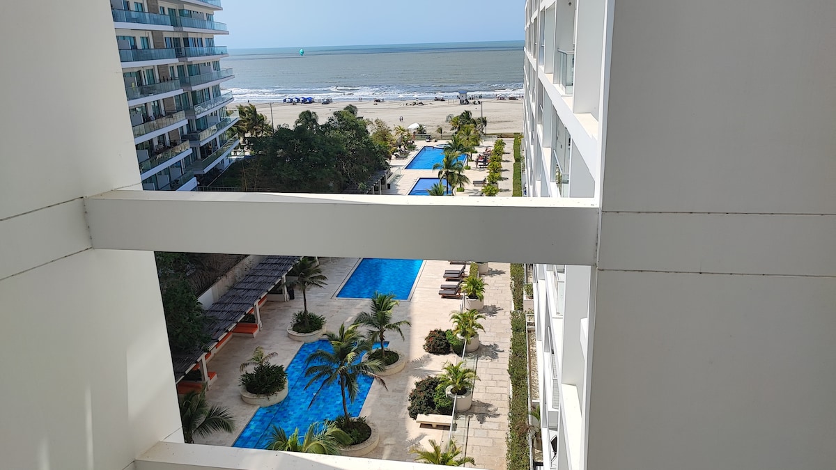 A view from the balcony showcases a well-maintained pool area surrounded by palm trees and lounge chairs. Beyond the pools, the sandy beach meets the ocean under a clear sky, highlighting a tranquil coastal atmosphere.