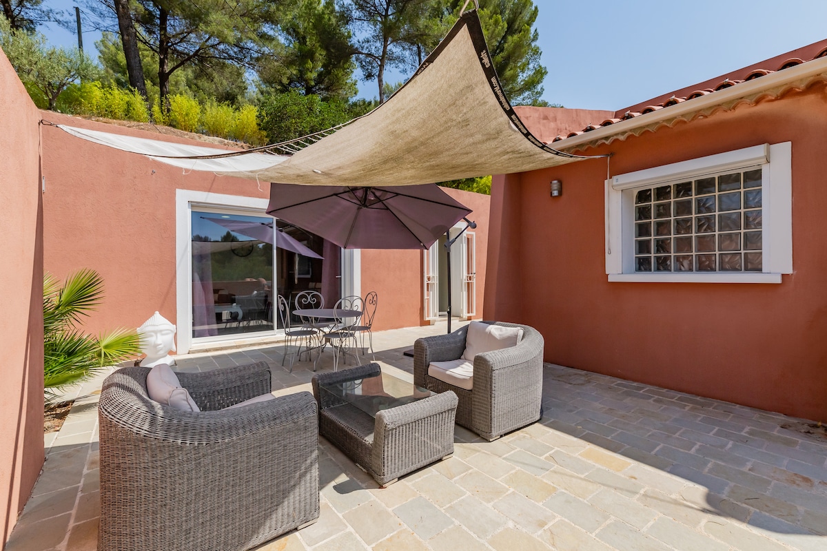 A private terrace is presented, featuring a seating arrangement with woven furniture. A central table is surrounded by four chairs, and a large umbrella provides shade. The area is bordered by greenery and has direct access to the residence through glass doors.