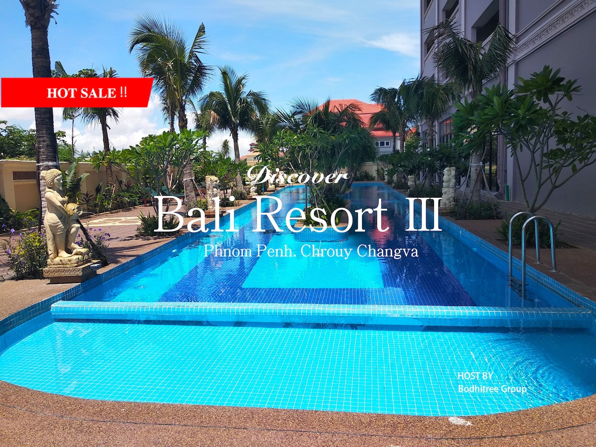 A long, serene swimming pool is visible, surrounded by tropical palm trees and manicured landscaping. The pool's turquoise water reflects the sunlight, with decorative statues positioned at either end. A modern building can be seen in the background, blending with the lush environment.