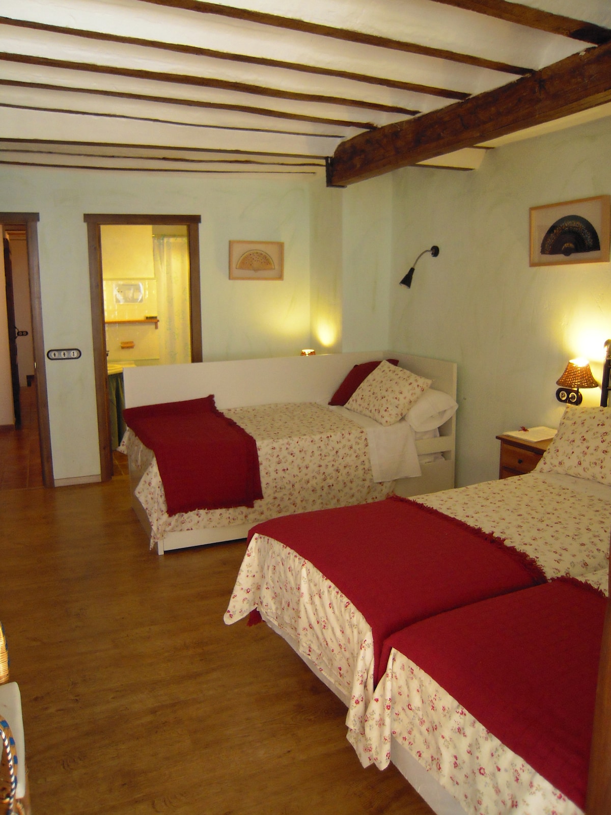 The room features two twin beds covered with patterned quilts in red and white, positioned against a light-colored wall. Wooden beams span the ceiling, complementing the wooden floor. Soft lighting illuminates the space, enhancing its warm atmosphere, while a doorway leads to a visible bathroom area.